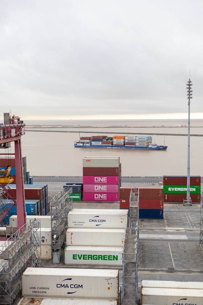 services-02 Containers stacked at a port with a cargo ship passing by on a cloudy day.