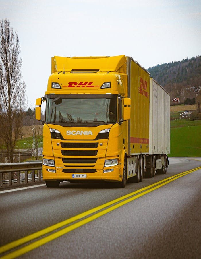 about-01 A yellow DHL truck on a scenic highway with green hills in the background.